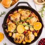 A skillet with roasted chicken thighs, potatoes, carrots, cranberries, and orange slices, garnished with rosemary. Rolls, orange slices, and cranberries are arranged around the pan.