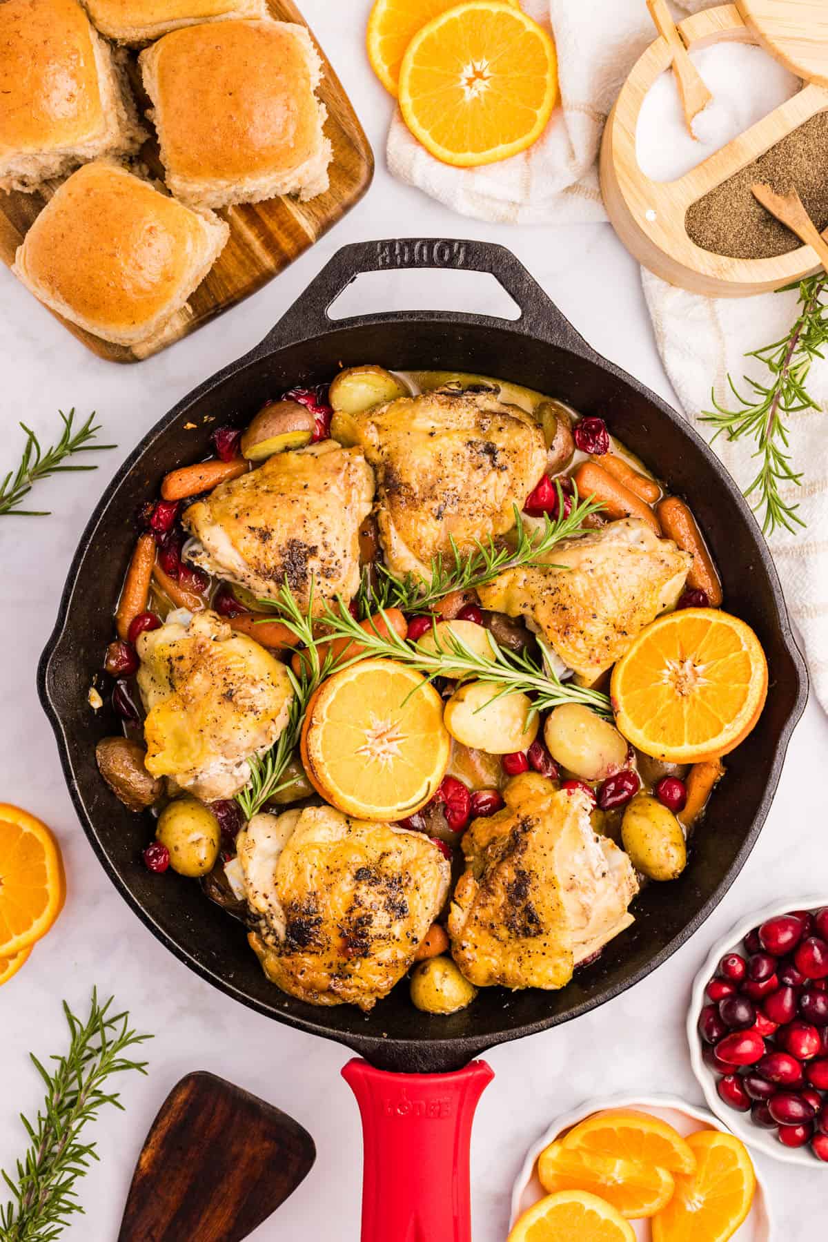 A skillet with roasted cranberry chicken thighs, potatoes, carrots, and orange slices, garnished with rosemary. Rolls, orange slices, and cranberries are arranged around the pan.