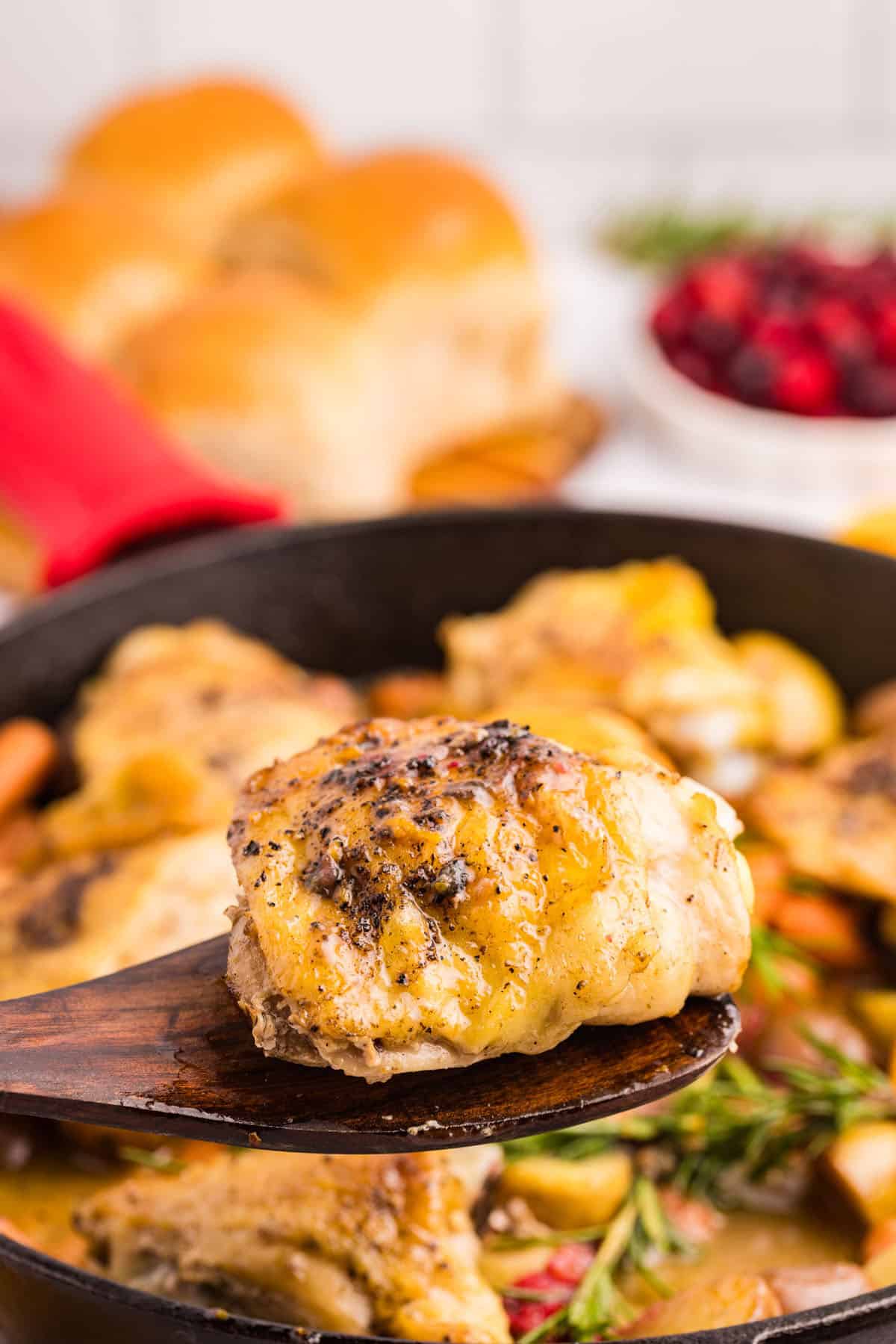 A wooden spoon holds a piece of seasoned, baked cranberry chicken above a skillet filled with more chicken. Bread rolls and a bowl of cranberries are visible in the background.