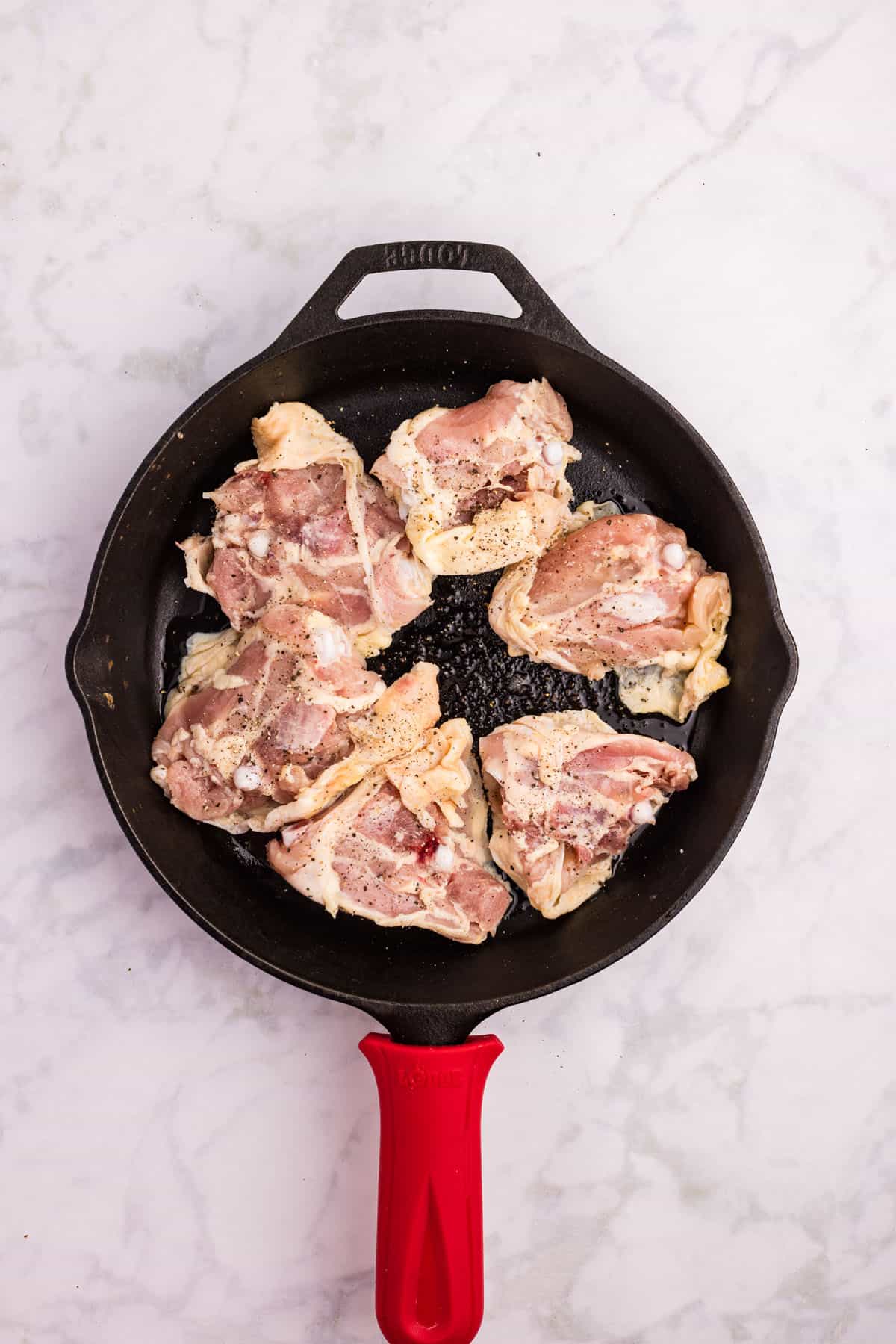 Raw chicken pieces seasoned with salt and pepper arranged in a black cast iron skillet with a red handle, on a white marble surface.