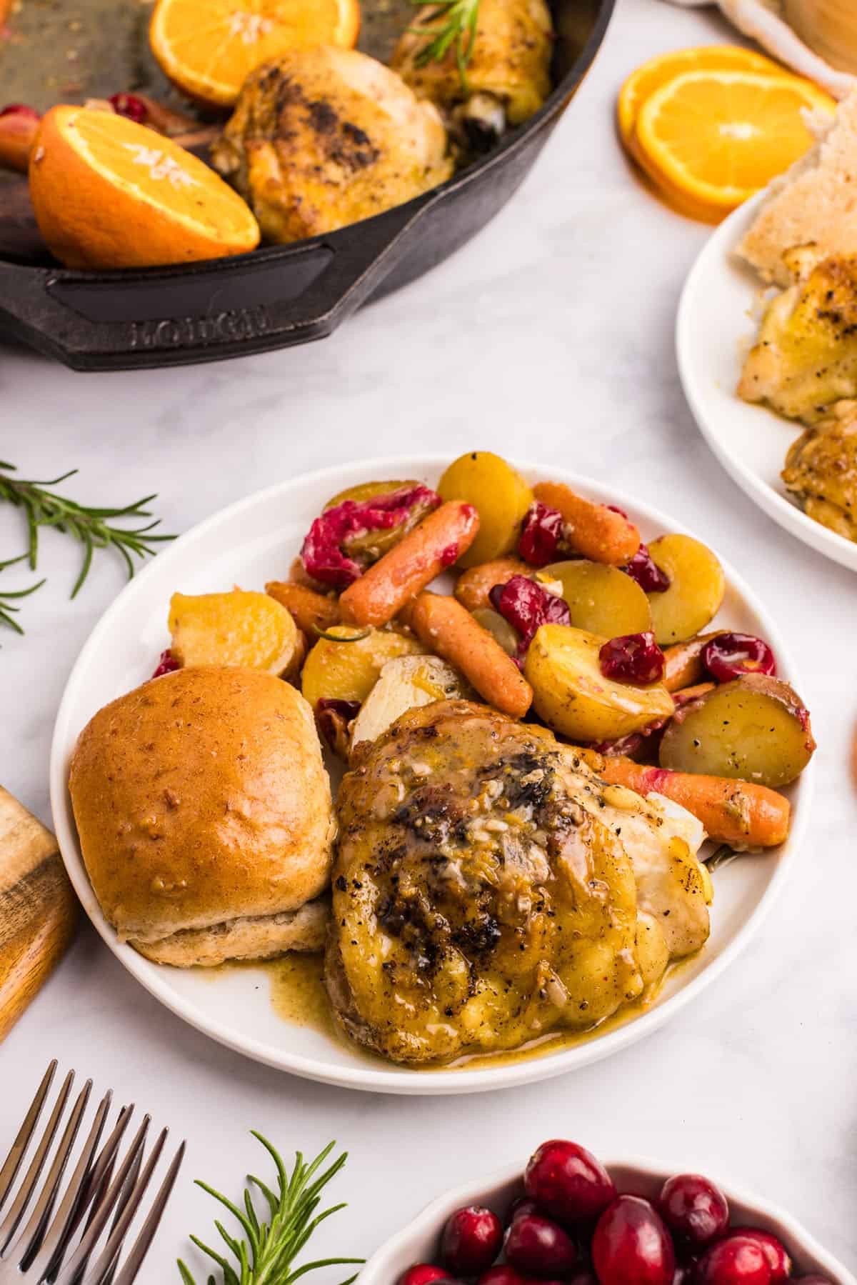 A plate with baked chicken, roasted potatoes, carrots, cranberries, and a dinner roll. A skillet with oranges and chicken in the background.