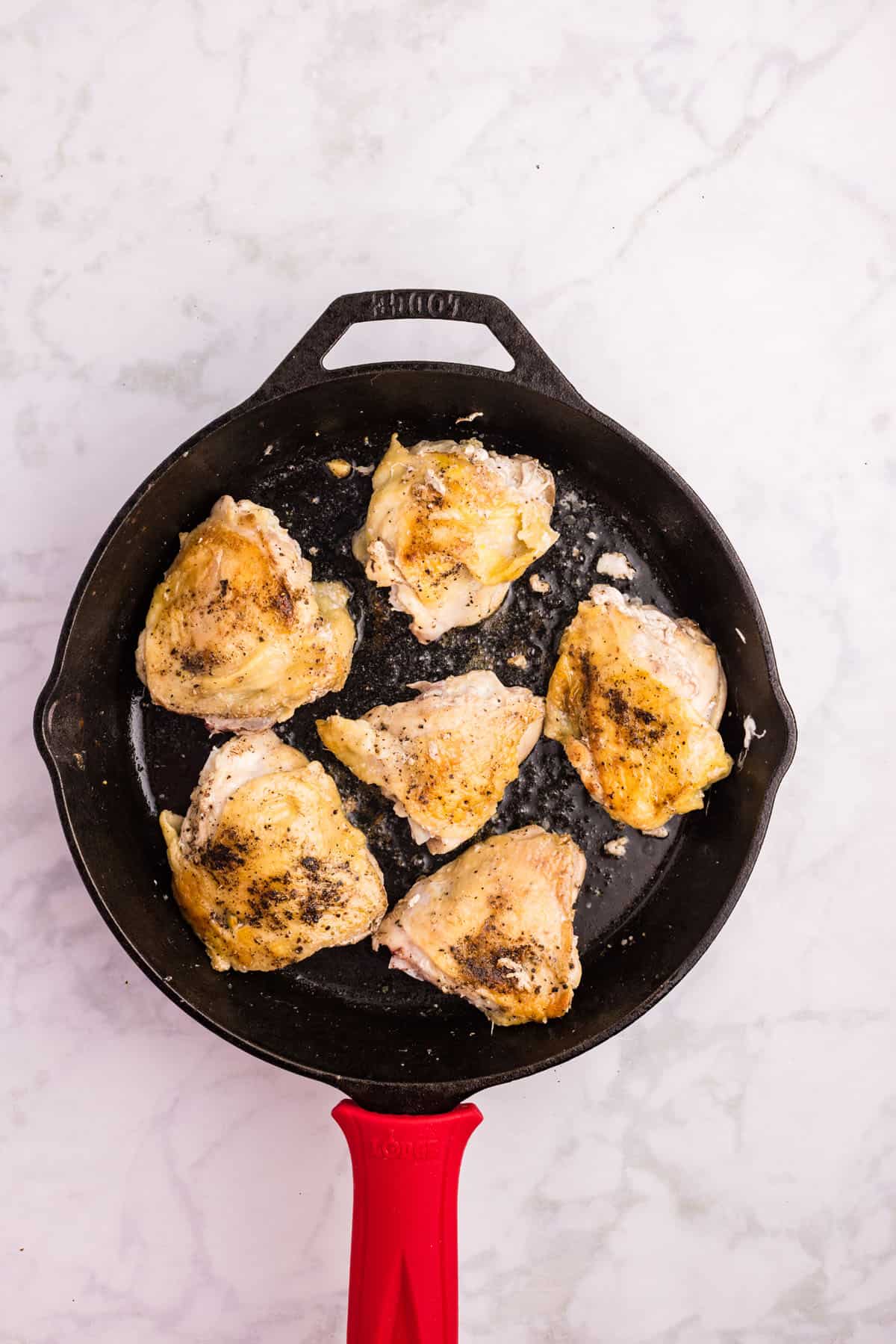 Six seasoned chicken thighs cooking in a black cast iron skillet with a red handle, shown on a white marble surface.