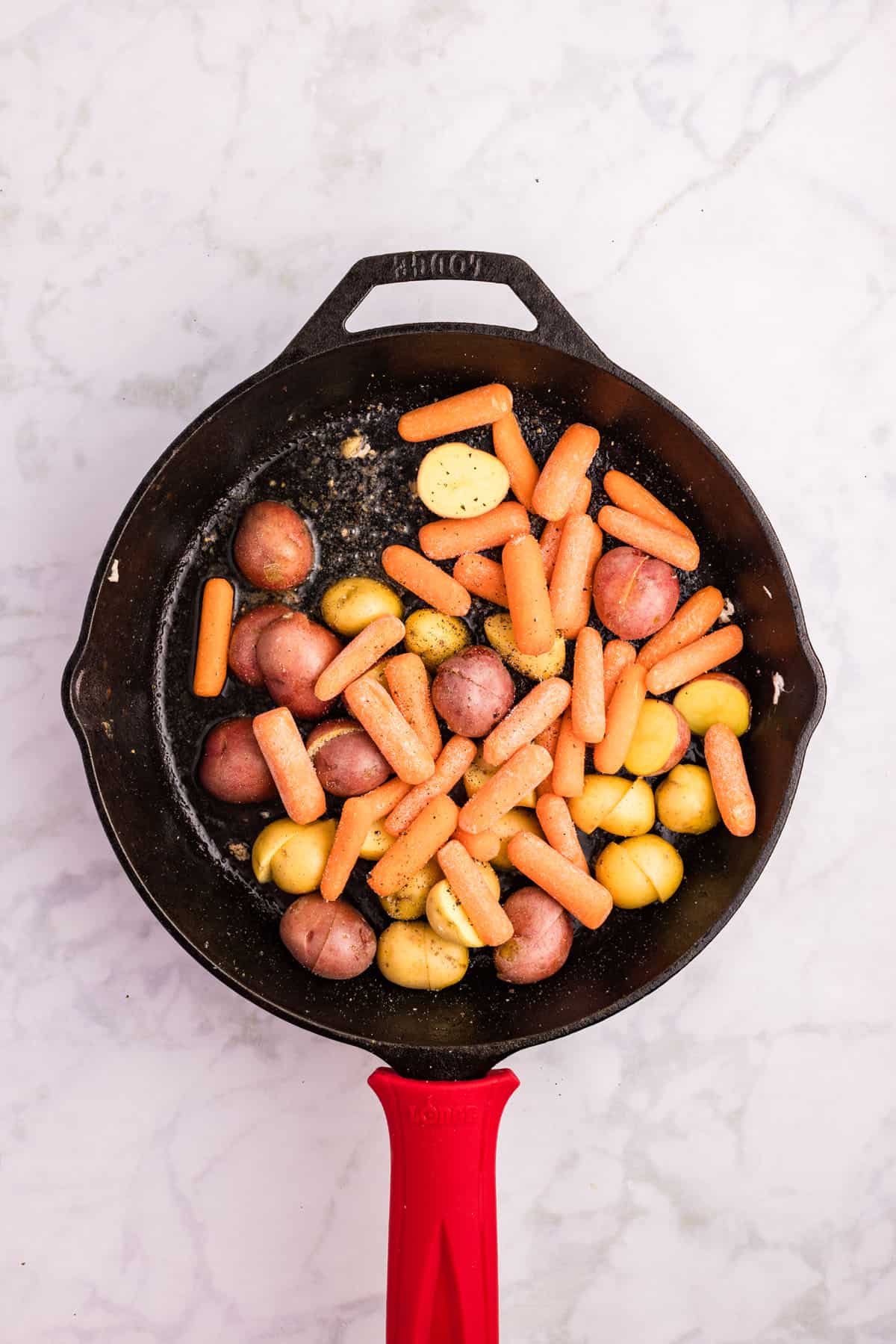 A black skillet with halved baby potatoes and baby carrots sits on a white marble surface, ready for cooking.