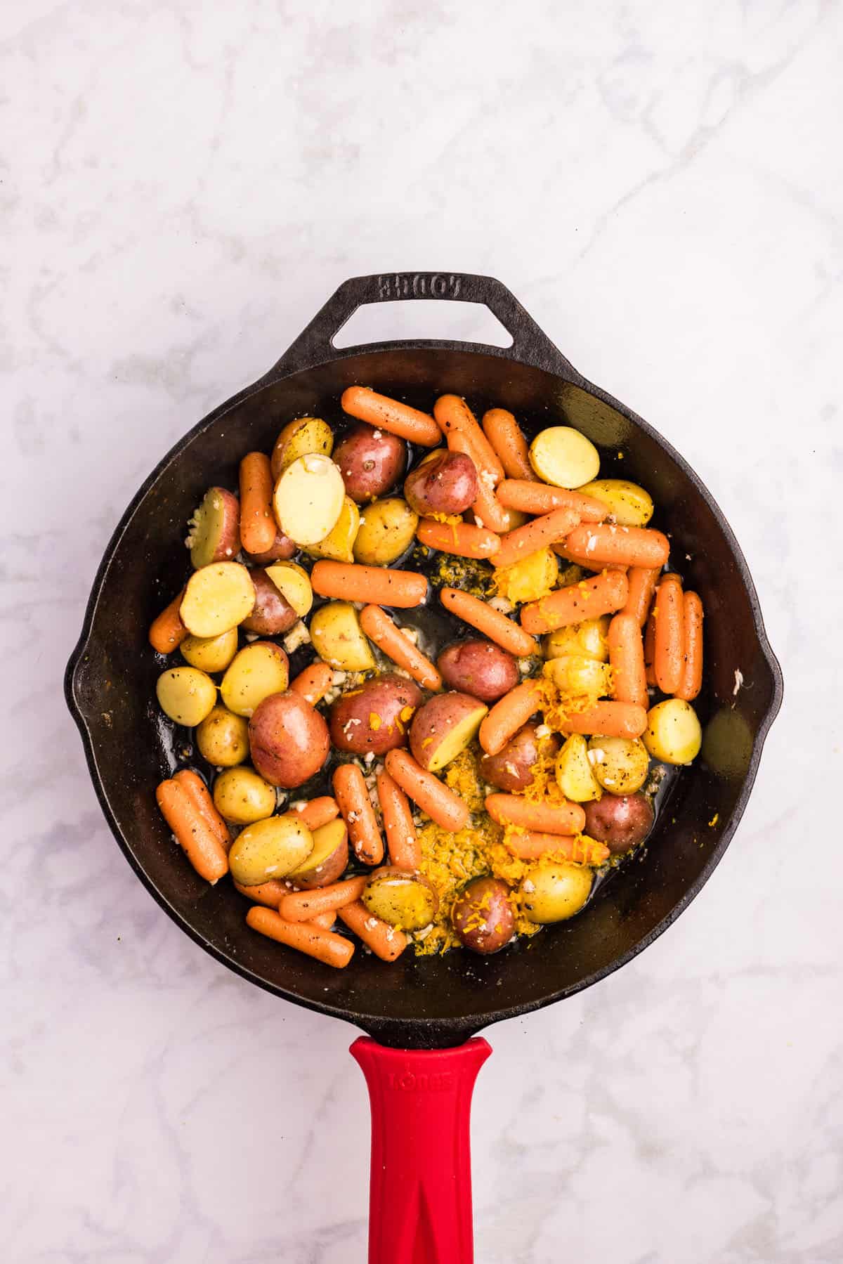 Cast iron skillet with roasted baby carrots and halved baby potatoes sits on a light marble surface.