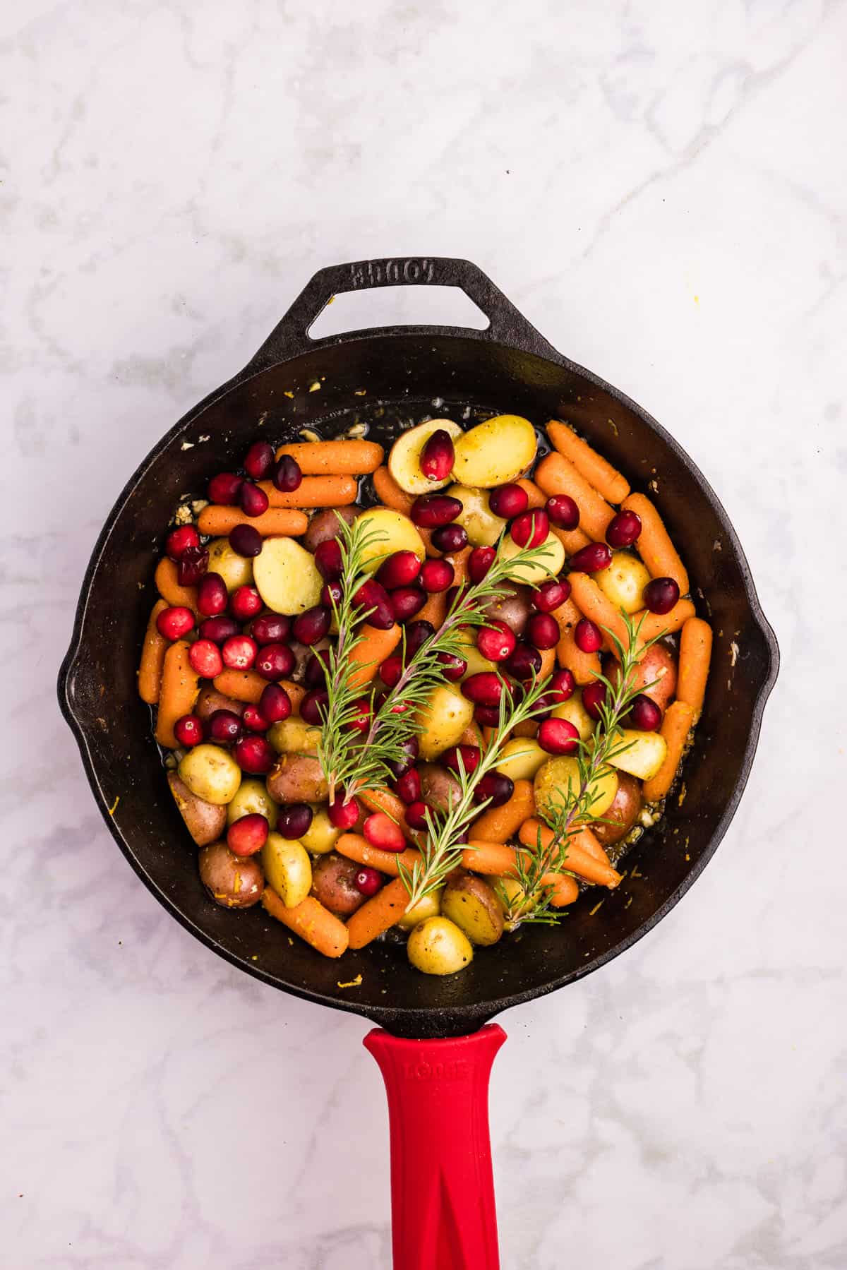 Cast iron skillet with baby carrots, halved small potatoes, cranberries, and sprigs of rosemary on a white marble surface.