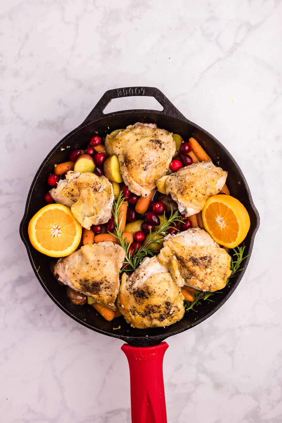 Cast iron skillet with roasted chicken thighs, carrots, potatoes, cranberries, fresh rosemary, and two orange halves on a marble surface.