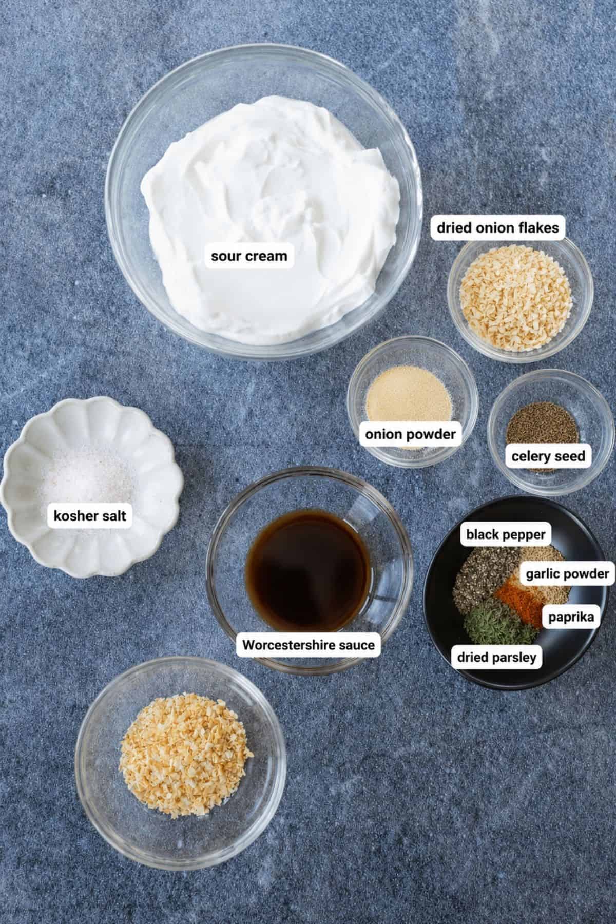 Bowls containing sour cream, kosher salt, Worcestershire sauce, dried onion flakes, onion powder, celery seed, black pepper, garlic powder, paprika, and dried parsley on a gray surface.