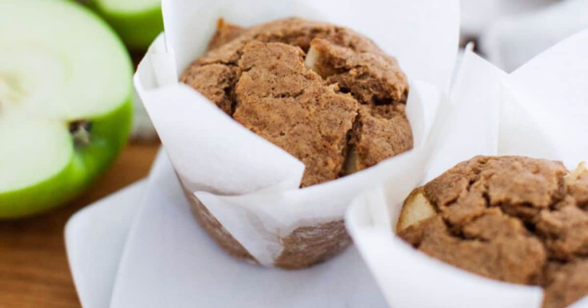 Two apple muffins in white parchment paper wrappers on a plate, with a green apple partially visible in the background.