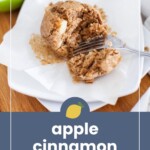 A partially eaten apple cinnamon muffin sits on a white plate with a fork, parchment paper, and a sliced green apple in the background.