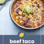 A bowl of beef taco soup topped with shredded cheese and avocado chunks, with part of a sliced avocado visible in the background.