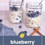 Two glass jars filled with overnight oats, milk, and blueberries, each with a spoon. Blueberries and two cinnamon sticks are on the table. Text reads “blueberry overnight oats.”.