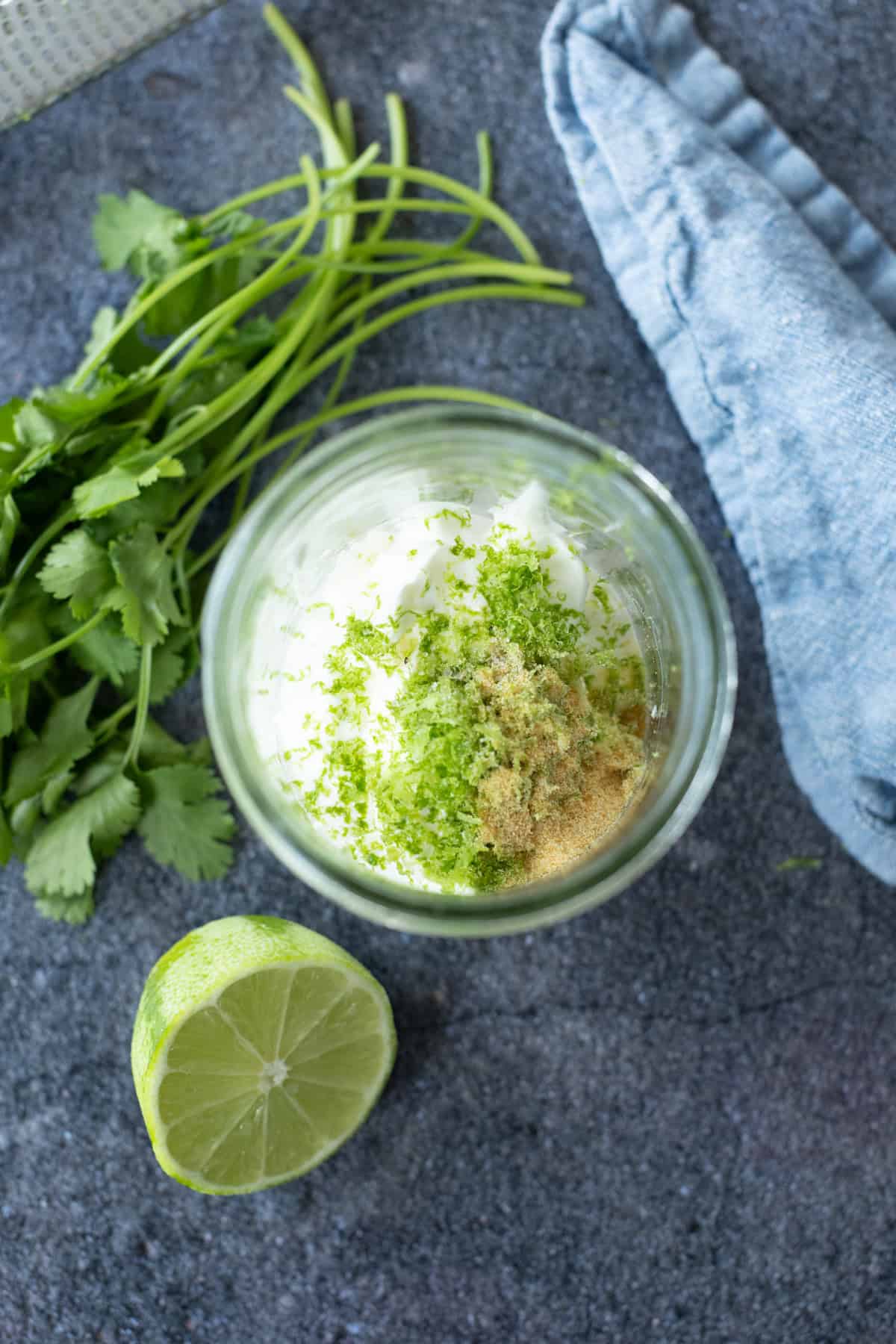 A glass jar with mayonnaise, lime zest, and spices sits on a gray counter beside fresh cilantro, a halved lime, and a folded blue cloth.