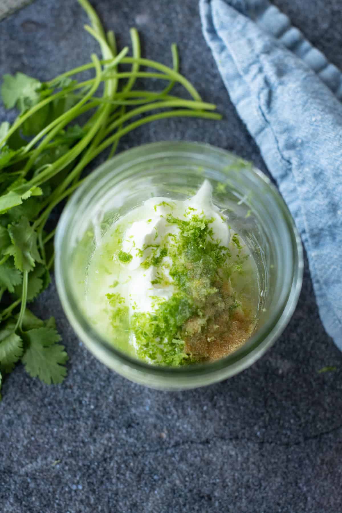 A glass jar with mayonnaise, lime zest, lime juice, and spices on a dark surface, next to fresh cilantro and a folded blue cloth.