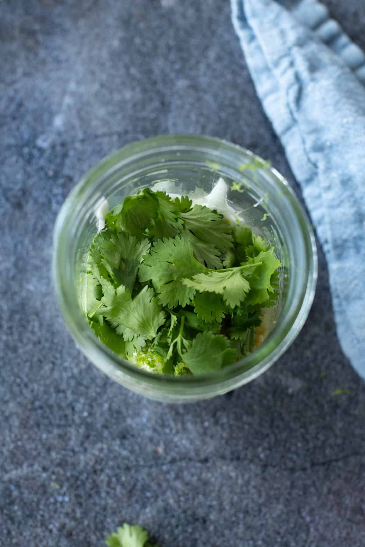 A glass jar filled with fresh cilantro leaves sits on a gray countertop next to a folded blue cloth.