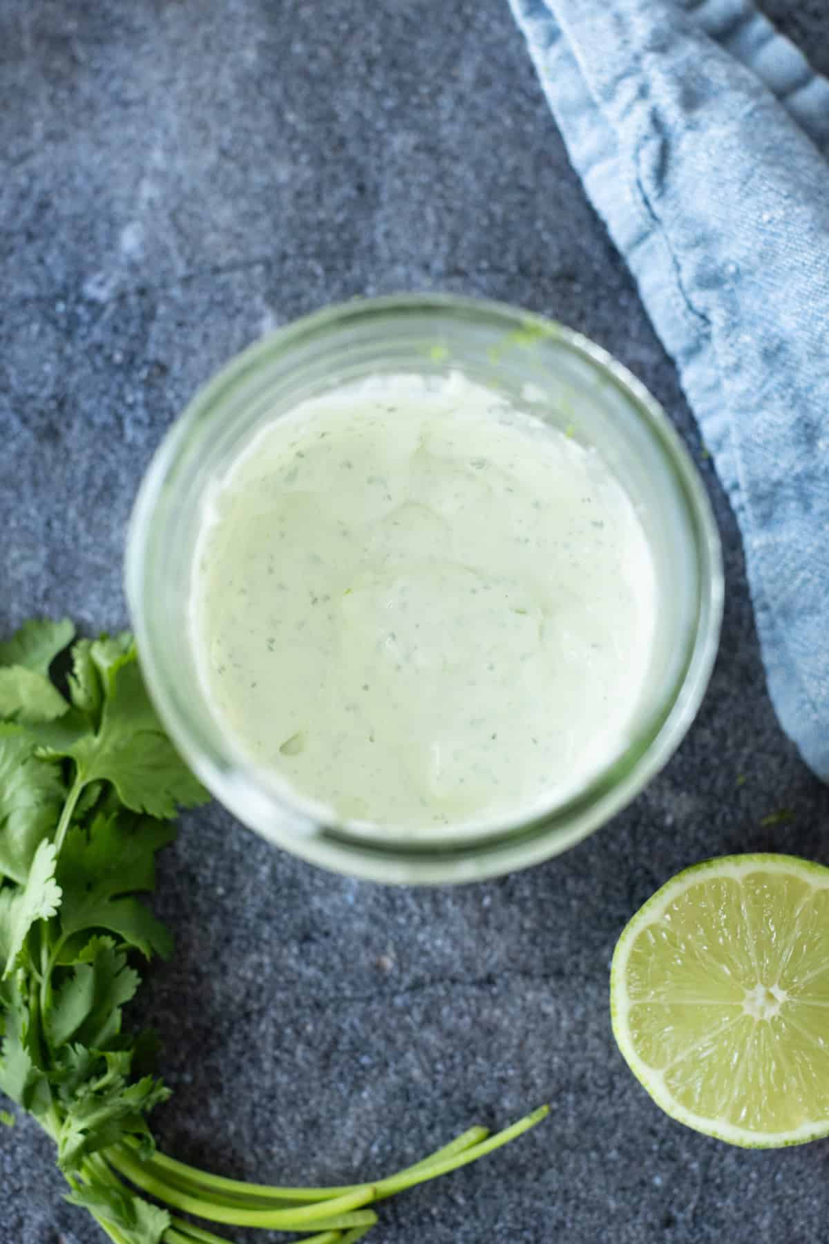 A glass jar of creamy green sauce sits on a gray surface, next to fresh cilantro, half a lime, and a blue cloth.