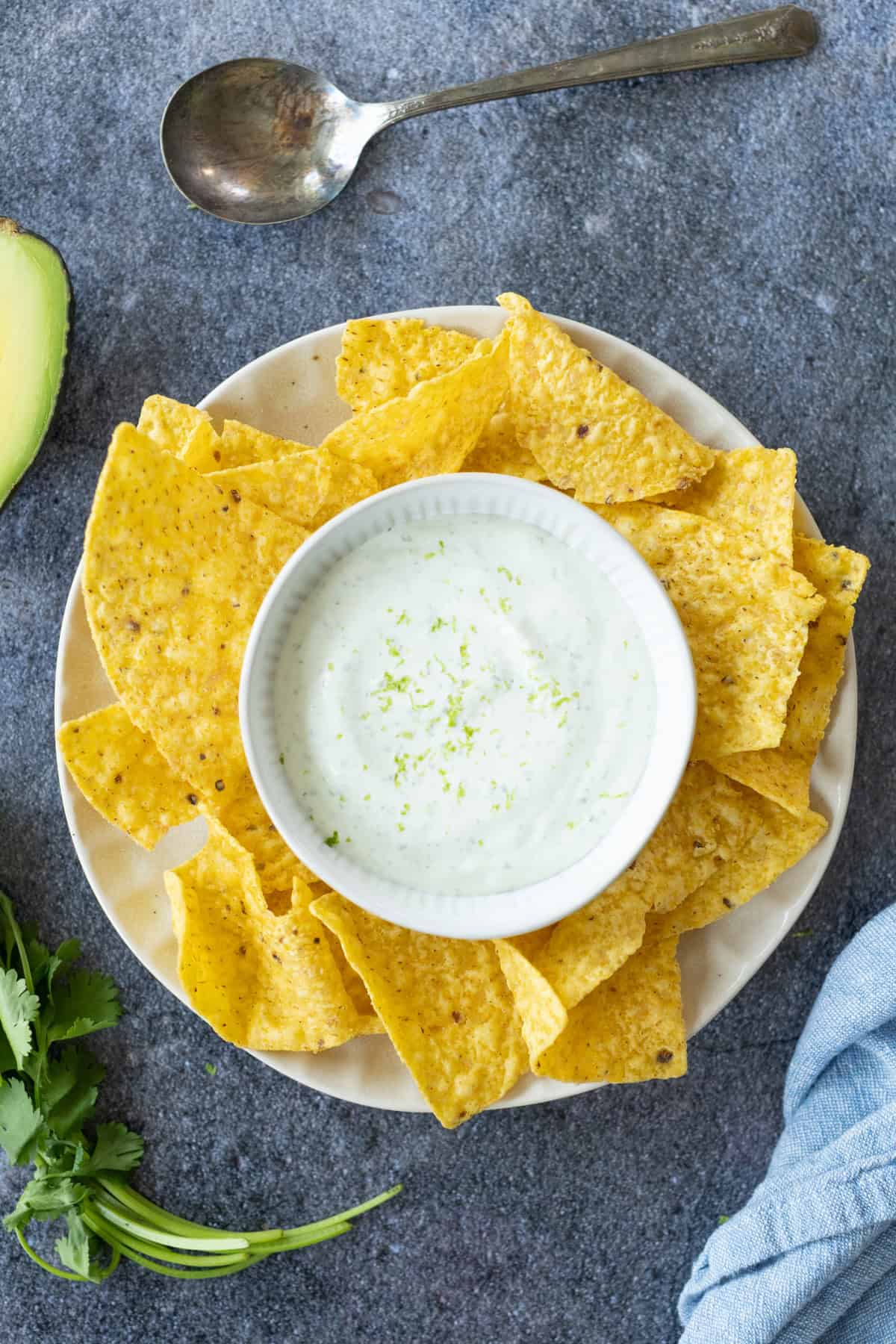 A plate of yellow tortilla chips surrounds a bowl of creamy cilantro lime crema garnished with lime zest. A spoon, cilantro, half an avocado, and a blue cloth are nearby on a gray surface.