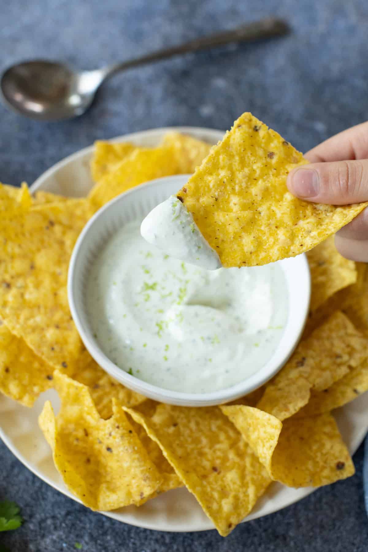 A hand dipping a tortilla chip into a bowl of creamy cilantro lime crema, surrounded by more chips on a plate. A spoon is in the background.