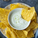 A plate of tortilla chips surrounds a bowl of creamy dip with lime zest on top; a spoon rests in the background.