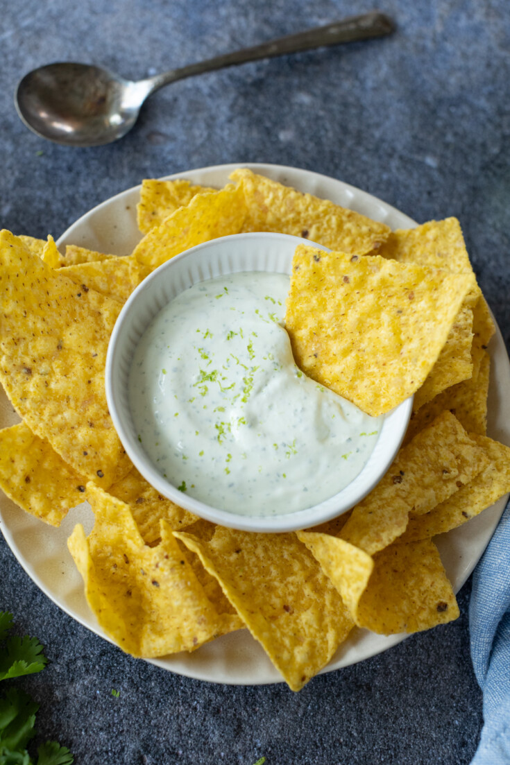 A plate of tortilla chips surrounds a bowl of creamy dip with lime zest on top; a spoon rests in the background.