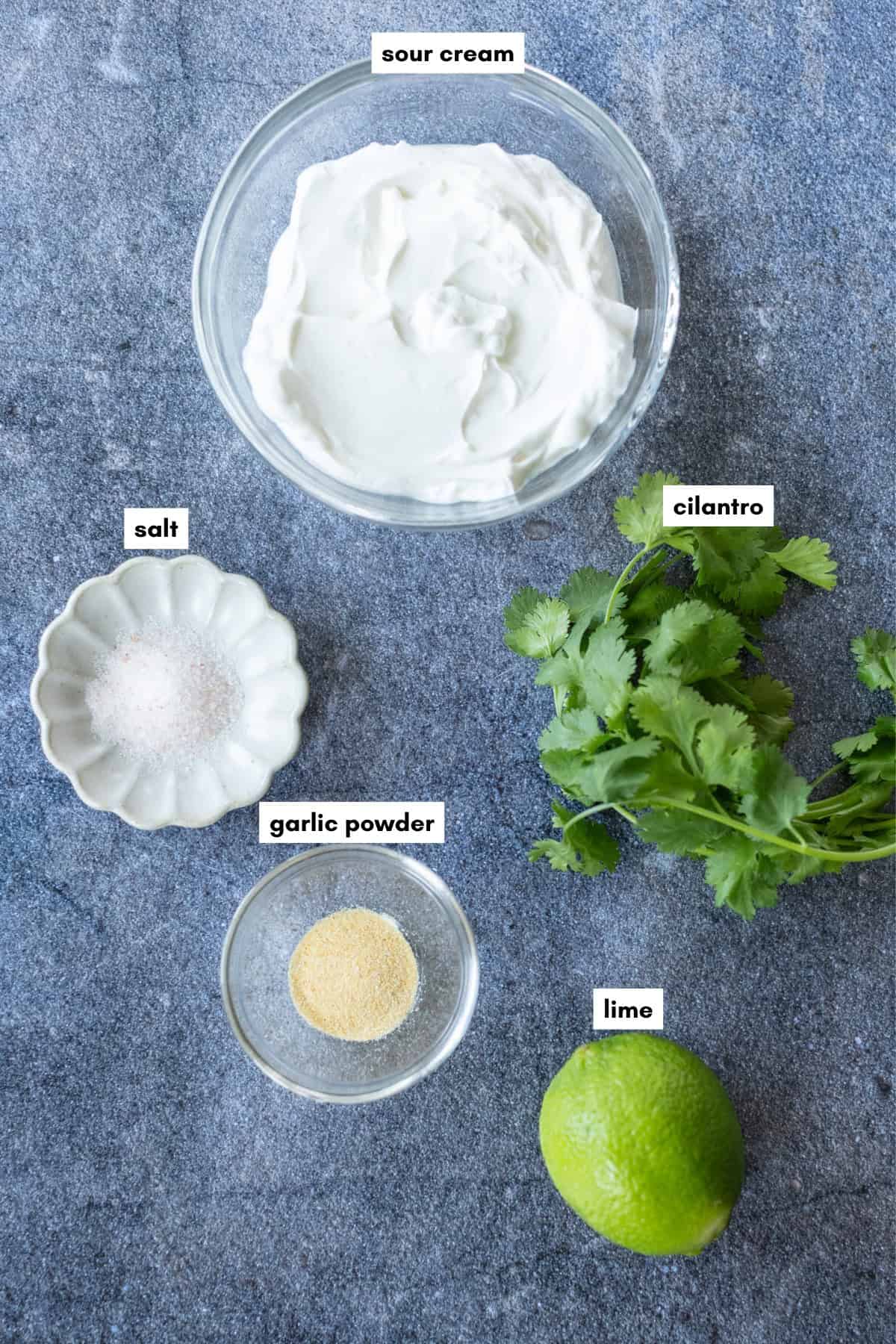 Glass bowls of sour cream and garlic powder, a small dish of salt, fresh cilantro, and a whole lime arranged on a blue surface. Labels identify each ingredient.