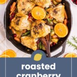 Skillet with roasted chicken thighs, small carrots, potatoes, cranberries, and orange slices, with a wooden spoon. Text reads “roasted cranberry chicken.”.