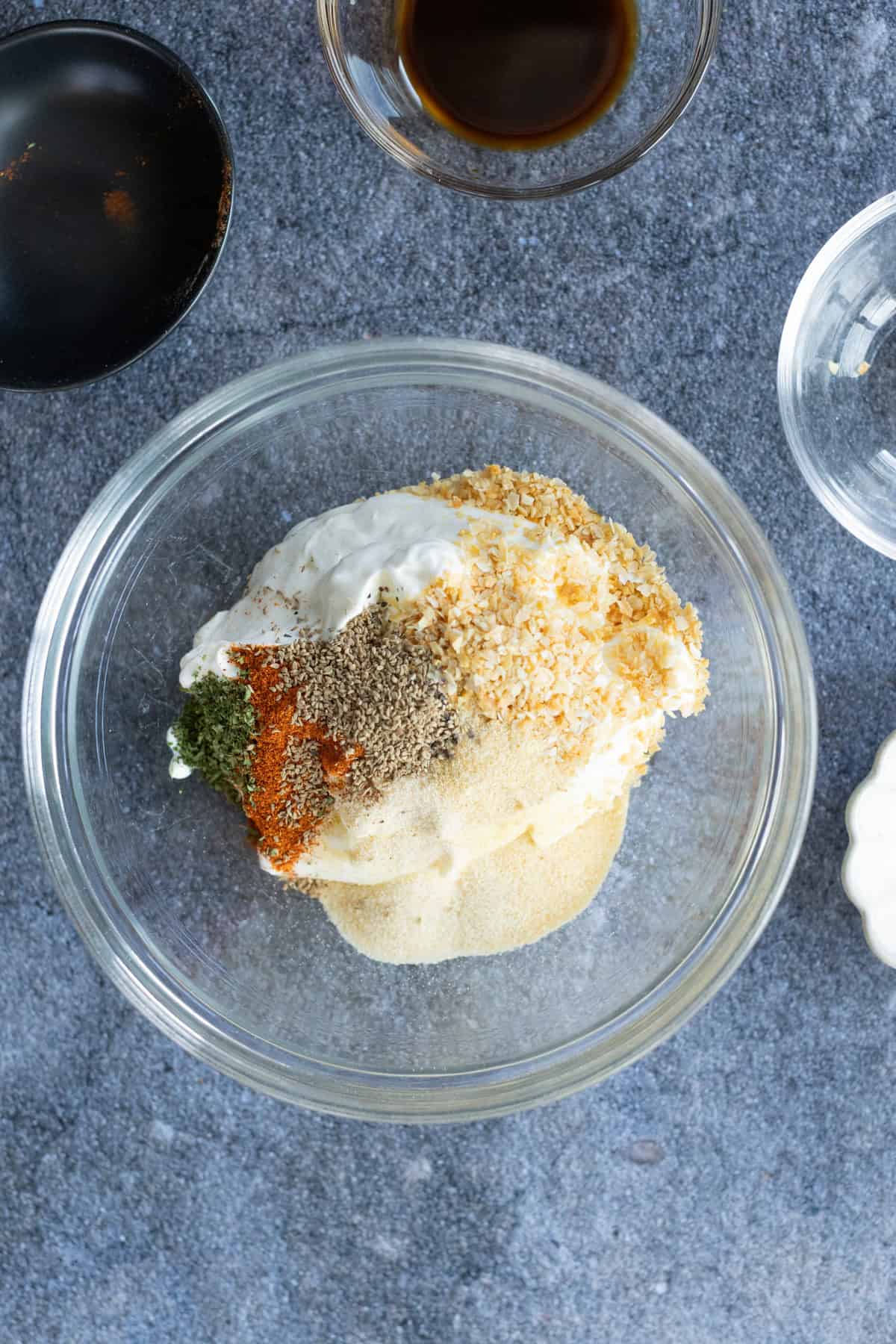 Clear glass bowl with sour cream, dried onion, onion powder, parsley, paprika, and other spices on a gray surface. Small bowls with liquid and seasoning are nearby.