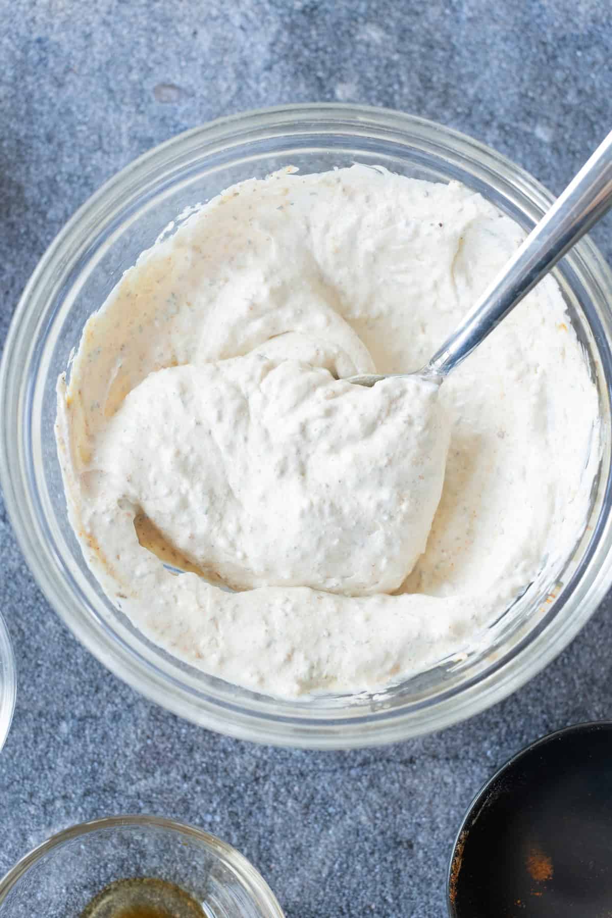 A glass bowl filled with a thick, creamy white mixture and a spoon resting inside, sitting on a gray surface.