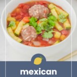 A bowl of Mexican meatball soup with meatballs, zucchini, tomatoes, carrots, and cilantro, placed on a light-colored surface.