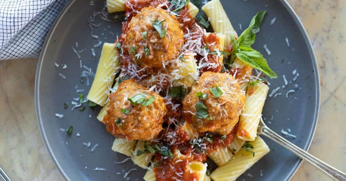 Three meatballs in tomato sauce on rigatoni pasta, garnished with grated cheese and basil, served on a gray plate with a fork.