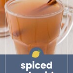 Clear glass cup filled with spiced apple cider, garnished with a cinnamon stick, placed on a saucer with another cup in the background.