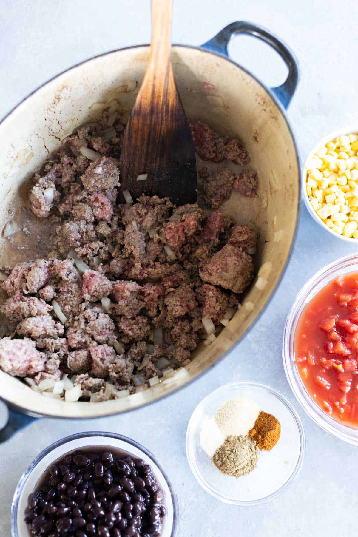 Ground beef and onions cooking in a pot, surrounded by bowls of black beans, corn, diced tomatoes, and seasonings.
