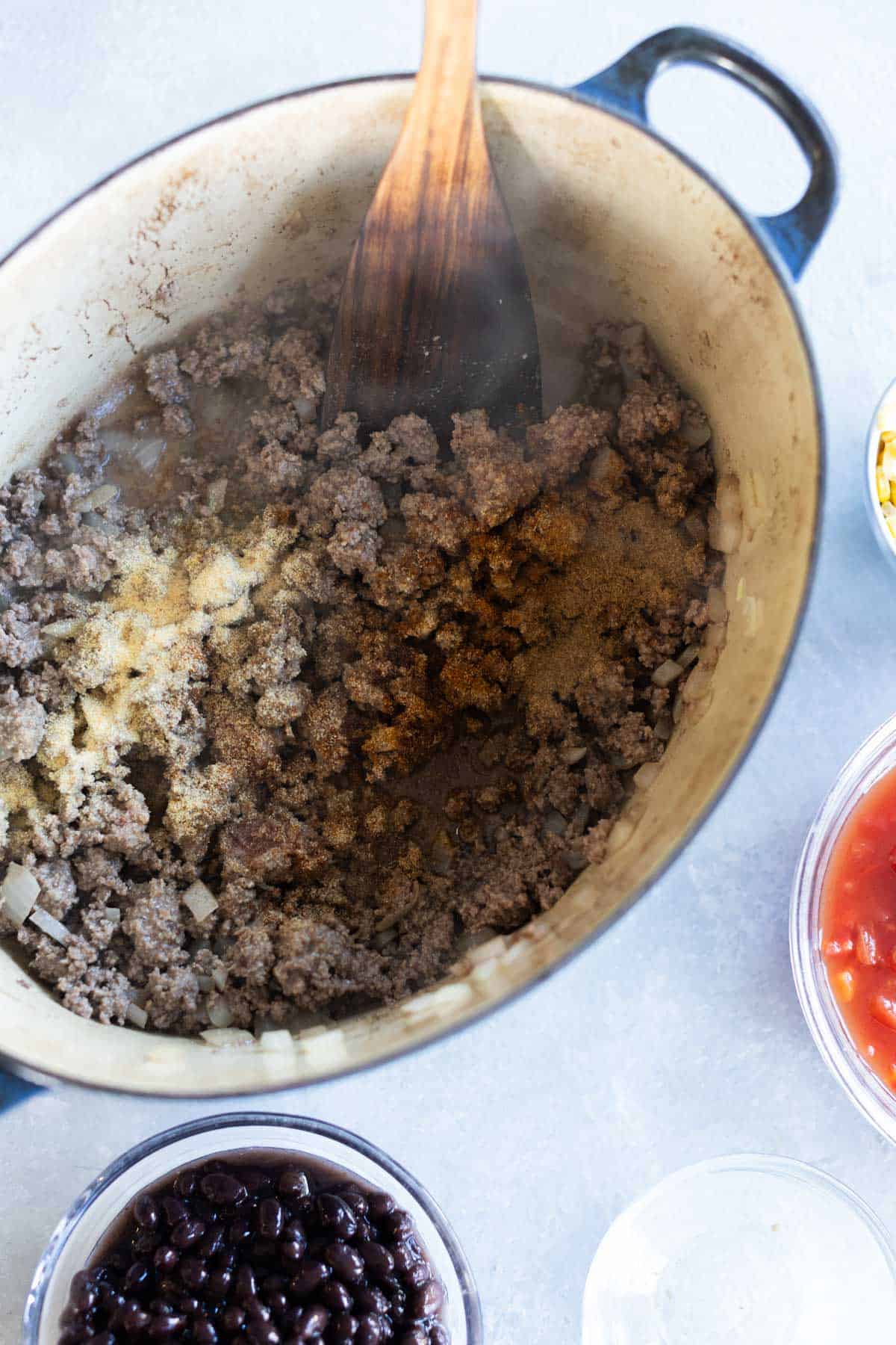 Ground beef is being cooked with seasonings in a large pot. Bowls of black beans, diced tomatoes, and corn are placed nearby on the counter.
