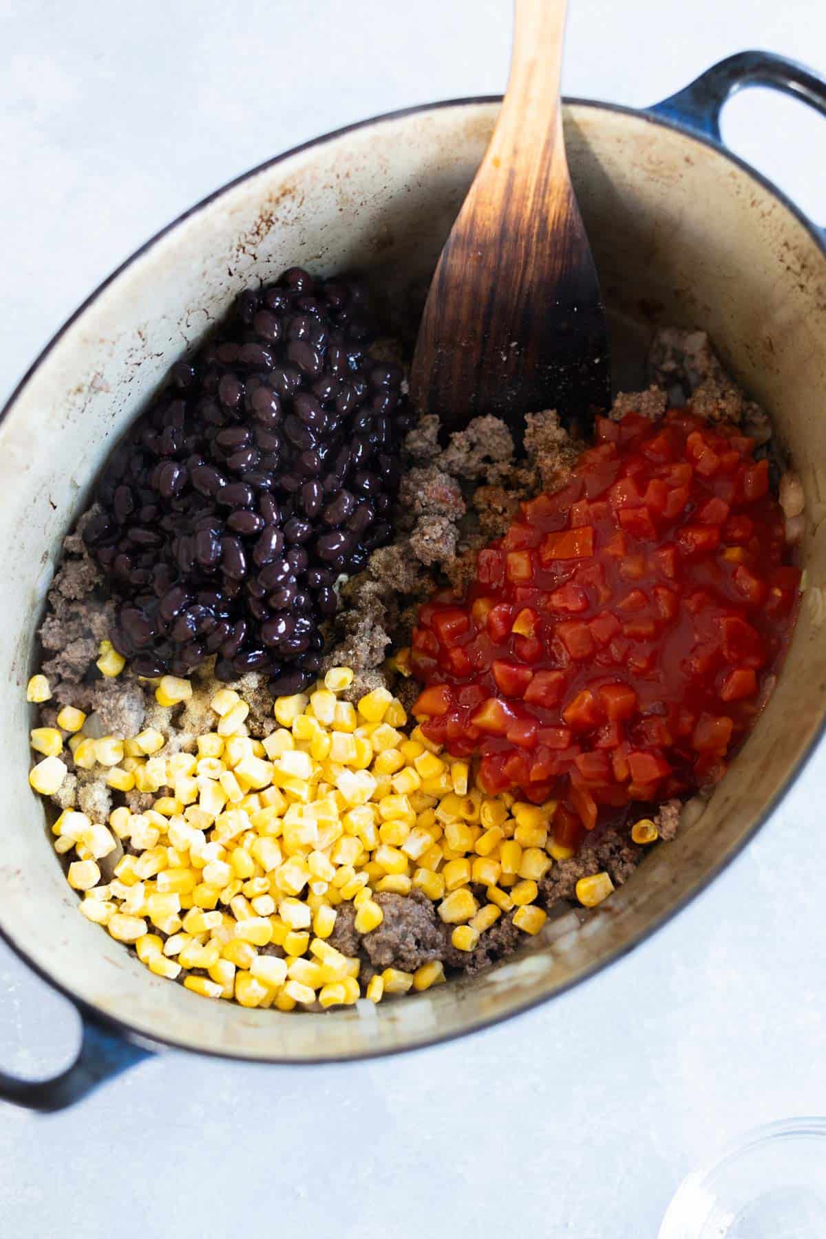 A pot with cooked ground beef, black beans, corn, and diced tomatoes, being mixed with a wooden spoon.