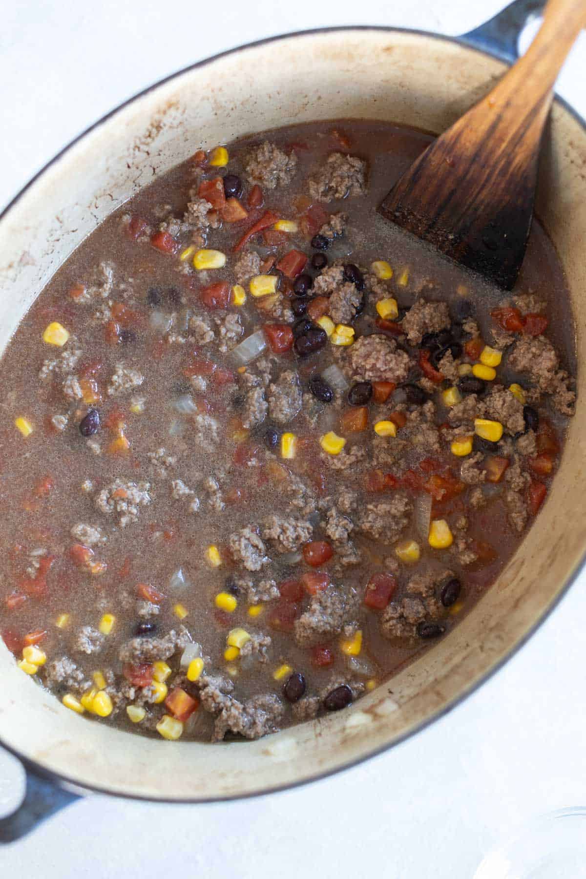 A pot of beef taco soup with ground beef, black beans, corn, diced tomatoes, and broth is being stirred with a wooden spoon.
