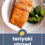 Plate of teriyaki-glazed salmon garnished with sesame seeds, served with broccoli and chopsticks on a white plate.
