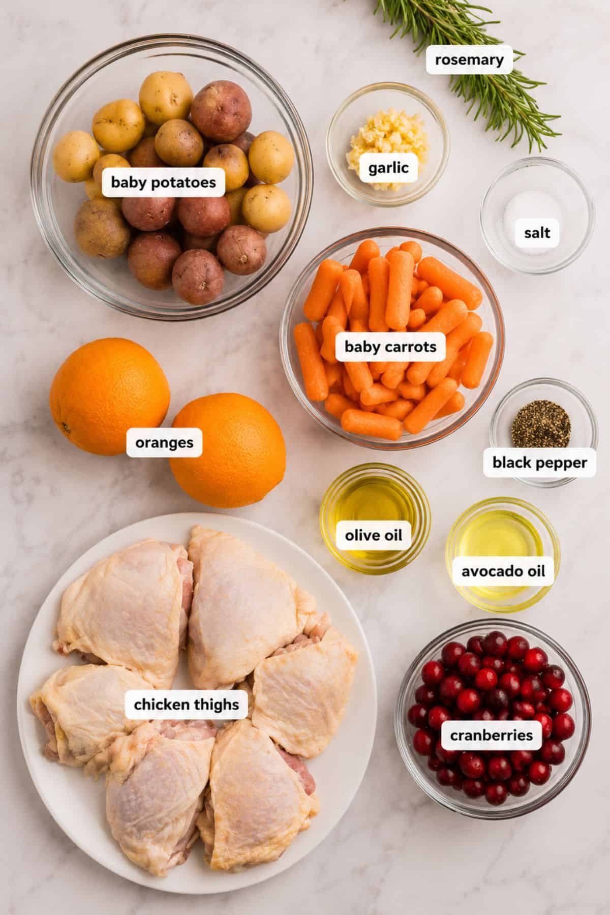 Ingredients on a counter: baby potatoes, baby carrots, cranberries, chicken thighs, oranges, rosemary, garlic, salt, black pepper, olive oil, and avocado oil—each labeled—promise a flavorful cranberry chicken recipe in the making.