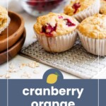 Cranberry orange muffins on a cooling rack, with a bowl of cranberries in the background and muffins on a wooden plate to the side.