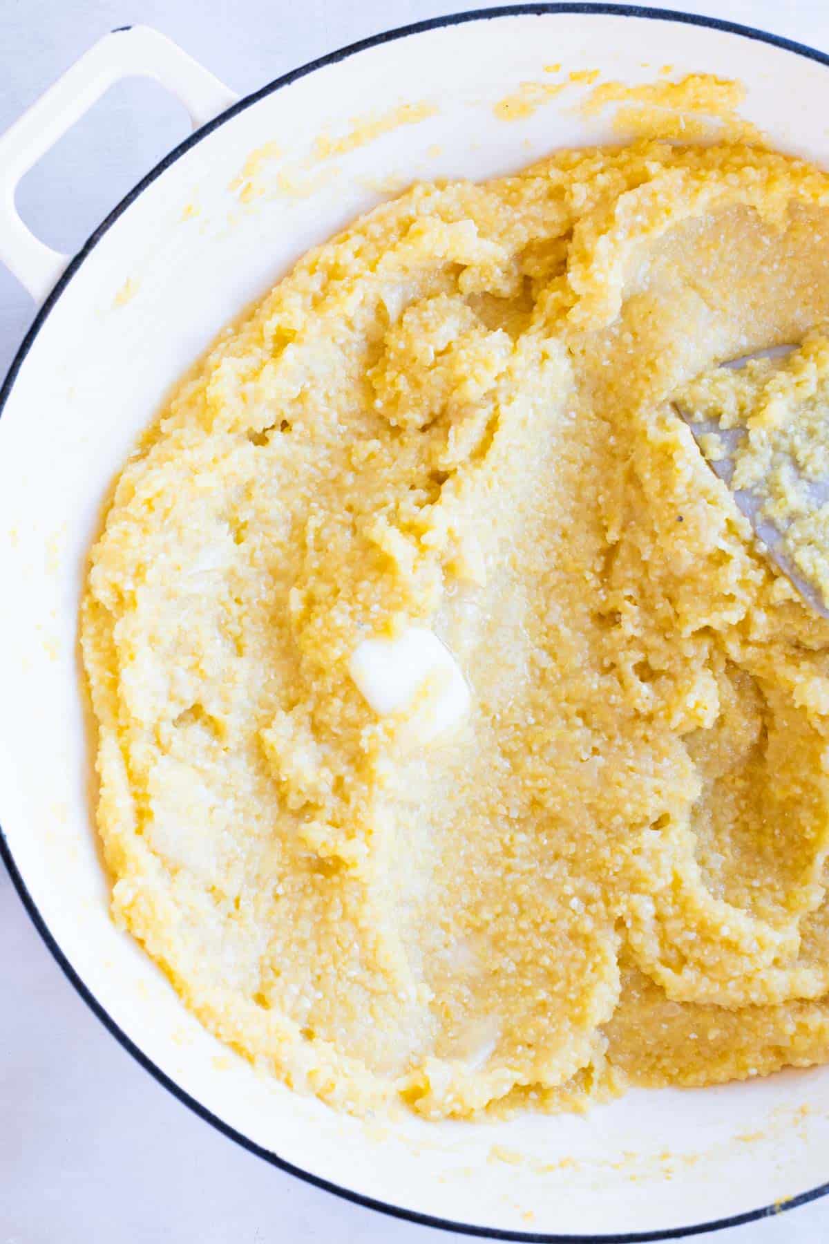 A white pot filled with creamy parmesan polenta, with a pat of butter melting on top and a spoon at the side.