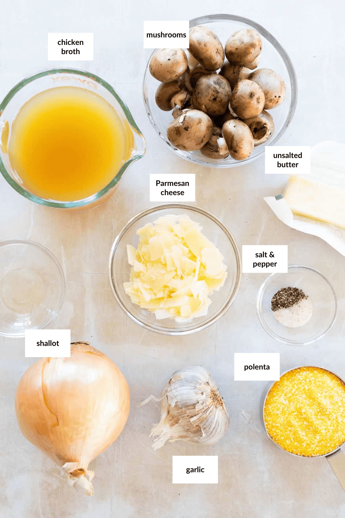 Ingredients for a side dish recipe arranged on a counter: chicken broth, mushrooms, unsalted butter, Parmesan cheese, salt, pepper, shallot, polenta, garlic, and an onion.