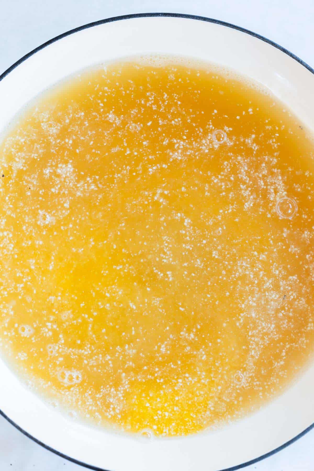 A close-up of a white bowl filled with yellow chicken broth, showing floating fat and bubbles on the surface.