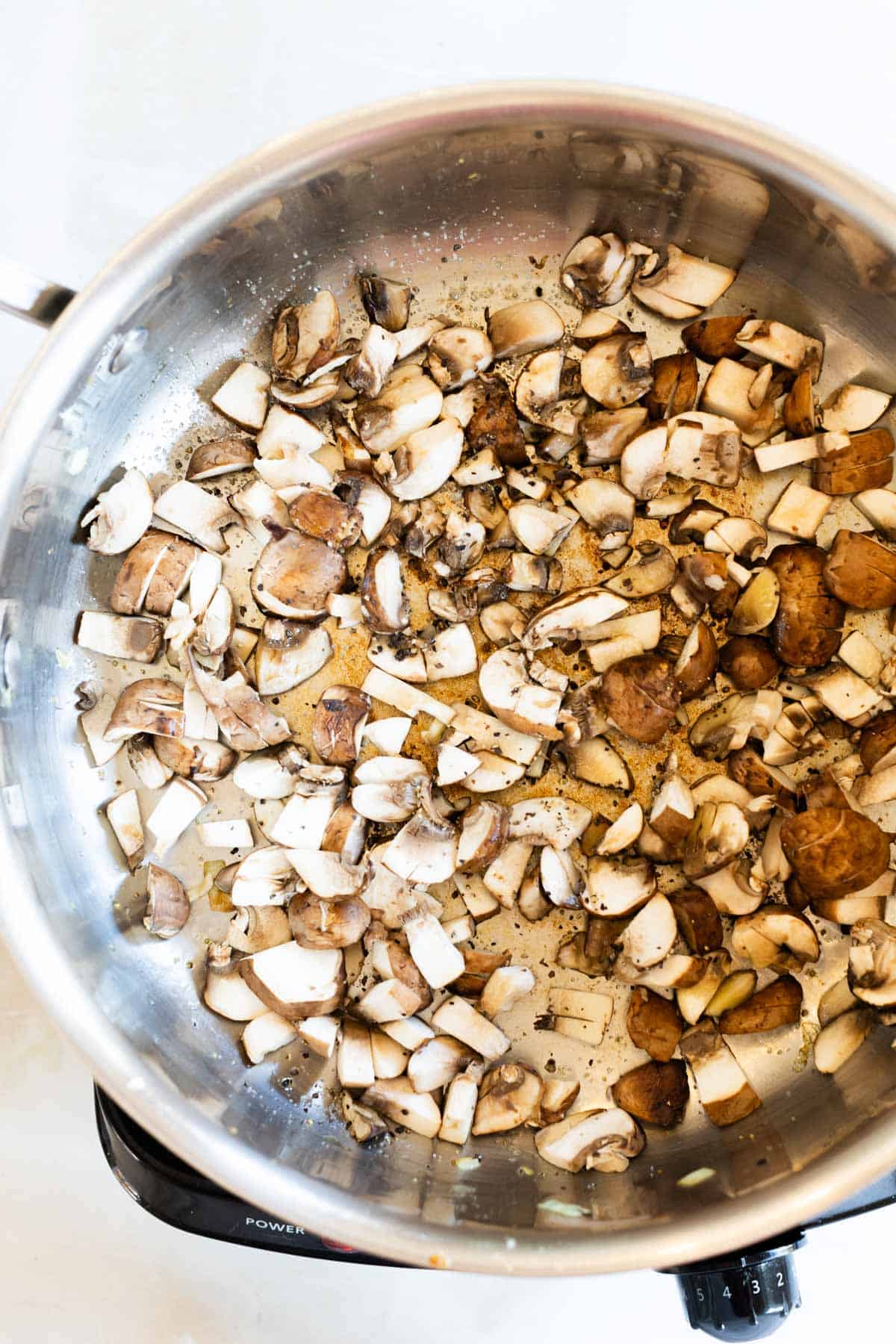 Chopped mushrooms seasoned with pepper cook in a stainless steel pan on a stovetop.