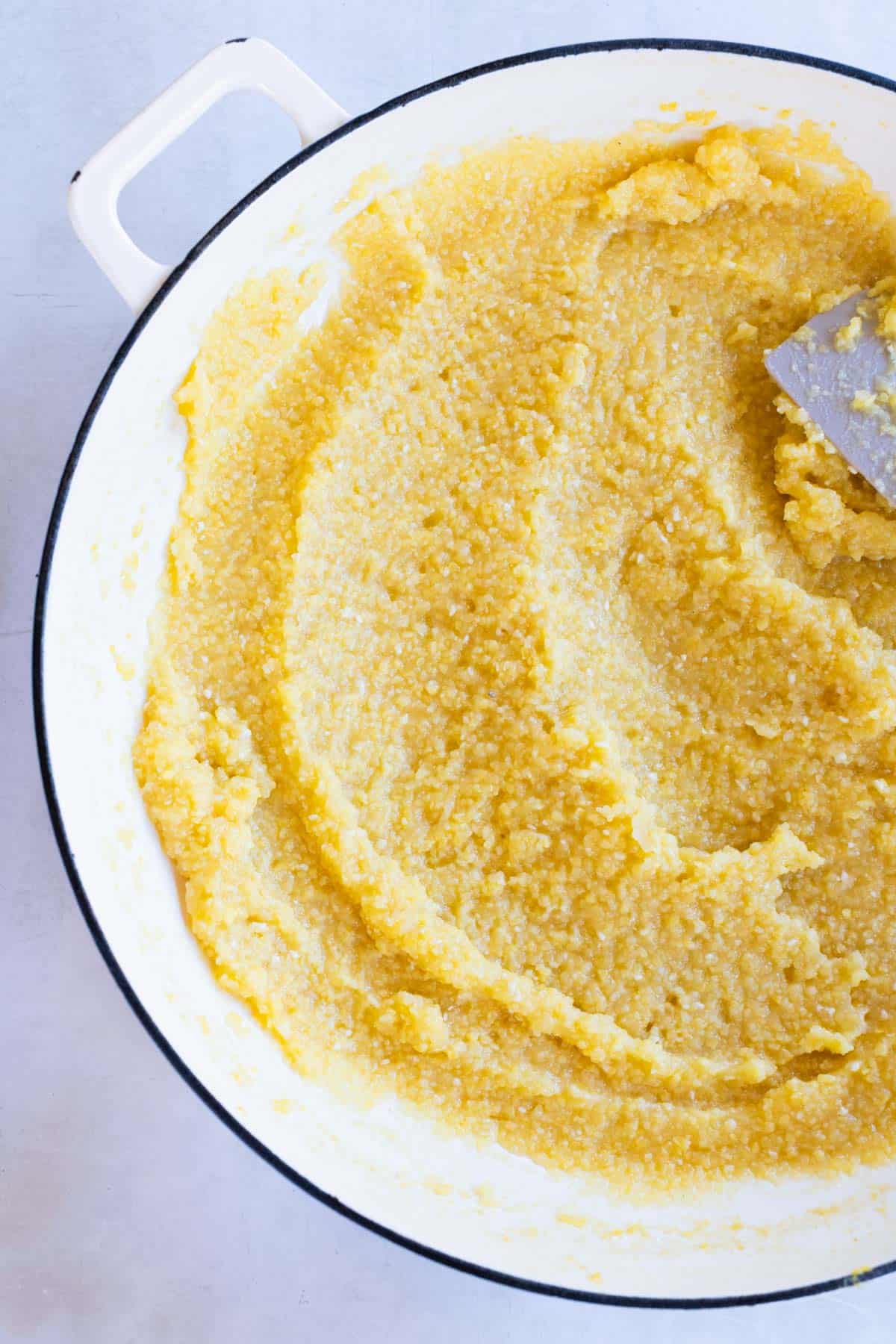 A white pot filled with cooked creamy polenta being stirred with a spatula.