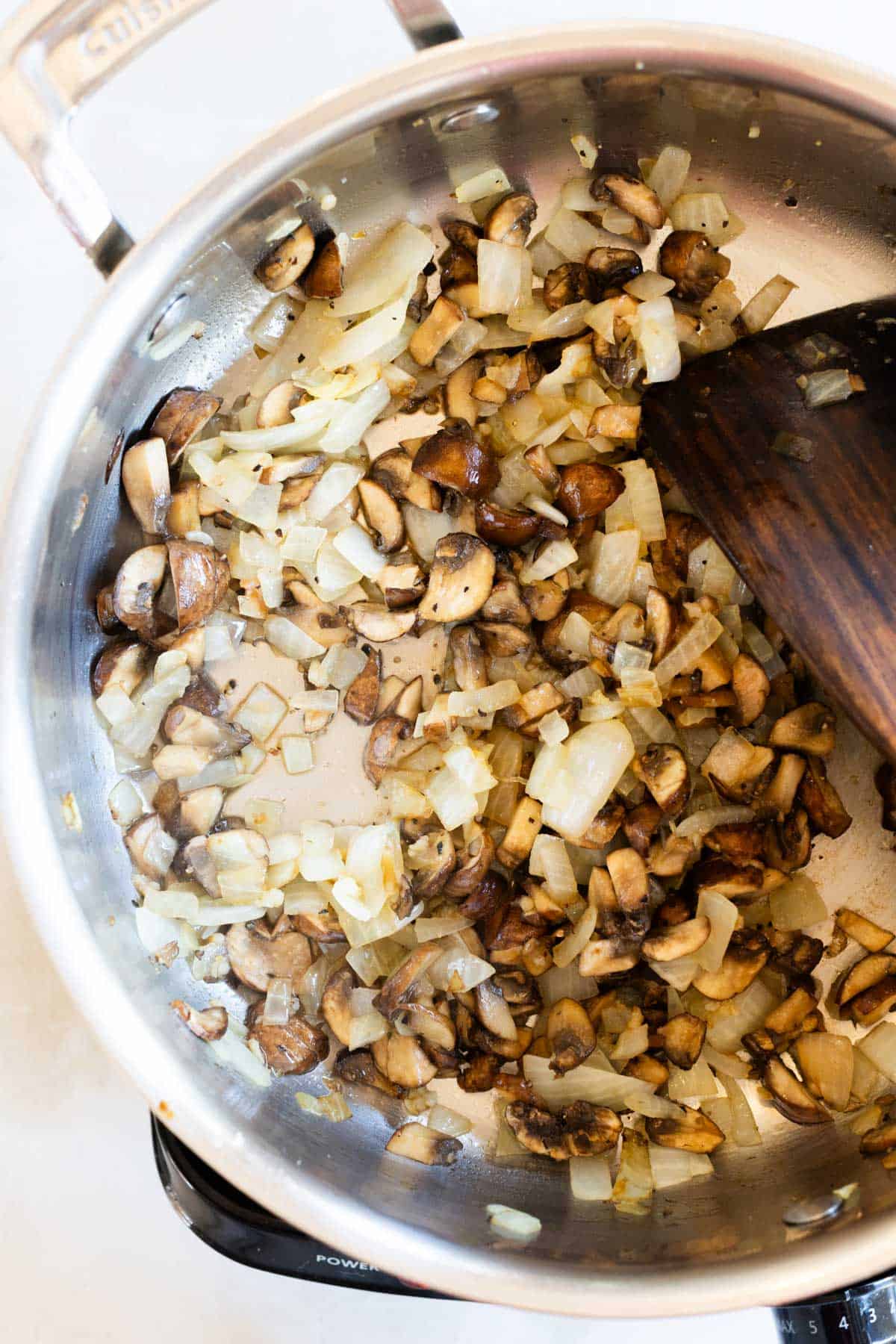 Diced onions and mushrooms are being sautéed in a stainless steel pan with a wooden spatula.