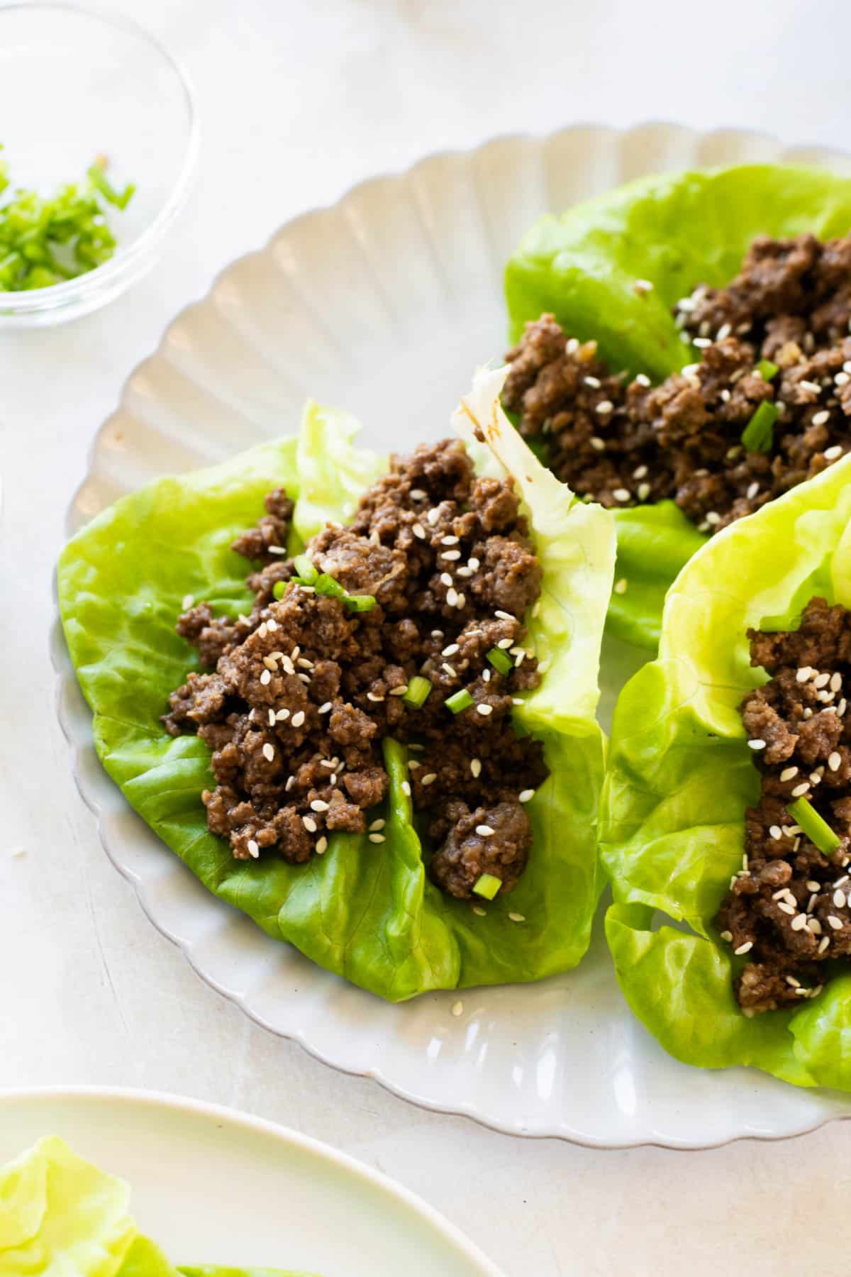 Three lettuce wraps filled with cooked ground beef, topped with sesame seeds and chopped green onions, arranged on a white plate.