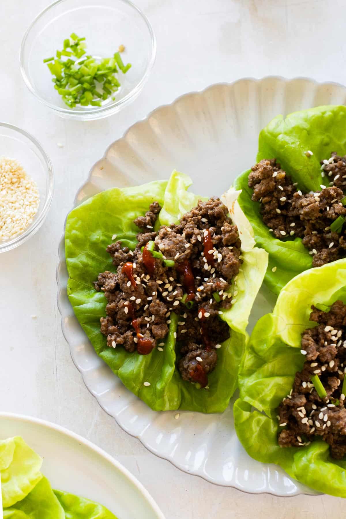 Three lettuce wraps filled with ground beef, topped with sesame seeds and sauce, are arranged on a plate. Nearby, small bowls contain chopped herbs and sesame seeds.