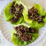 Three Asian ground beef lettuce wraps filled with ground meat mixture and sprinkled with sesame seeds are arranged on a white plate.