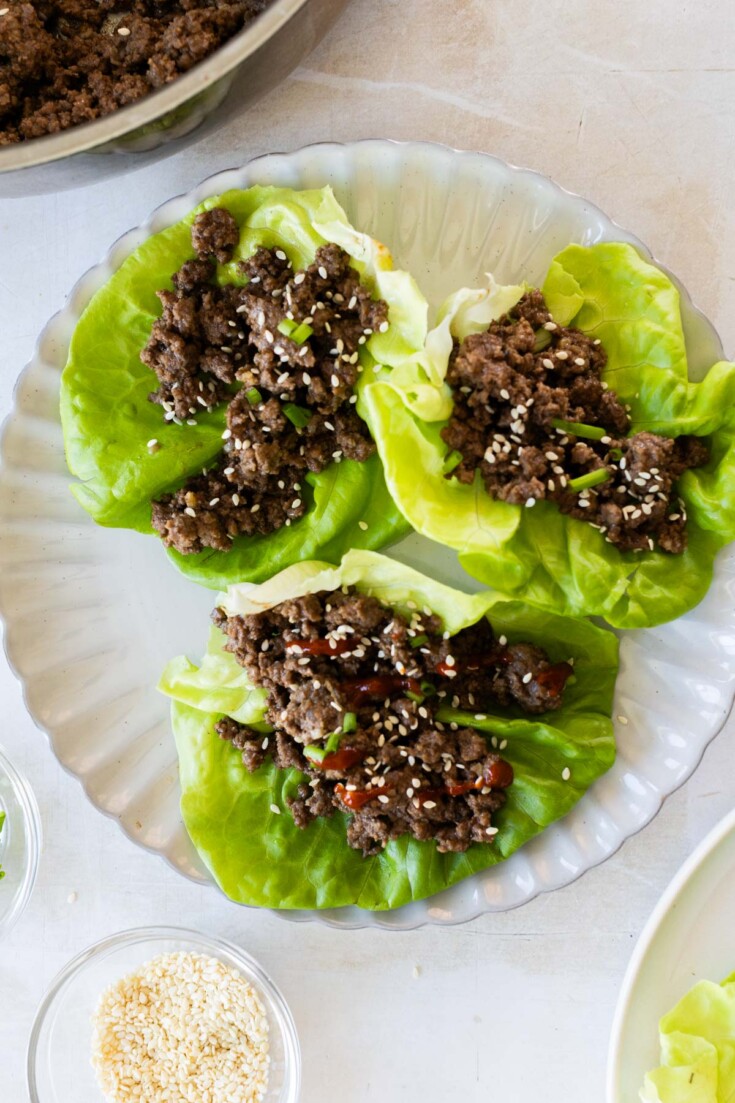 Three Asian ground beef lettuce wraps filled with ground meat mixture and sprinkled with sesame seeds are arranged on a white plate.