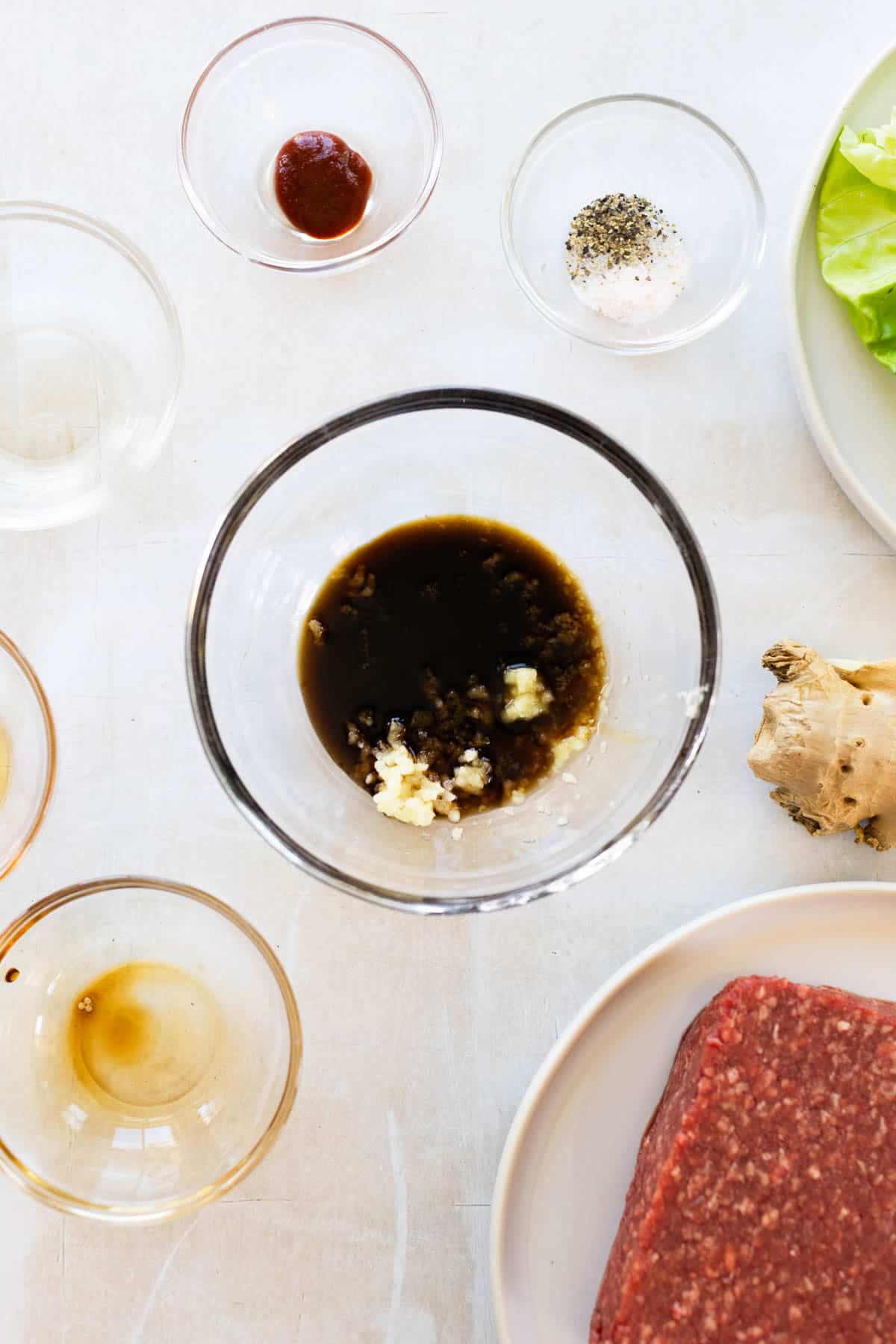 Ingredients for a recipe including ground beef, soy sauce with garlic, seasonings, a ginger root, lettuce leaves, and small bowls of sauces on a white surface.
