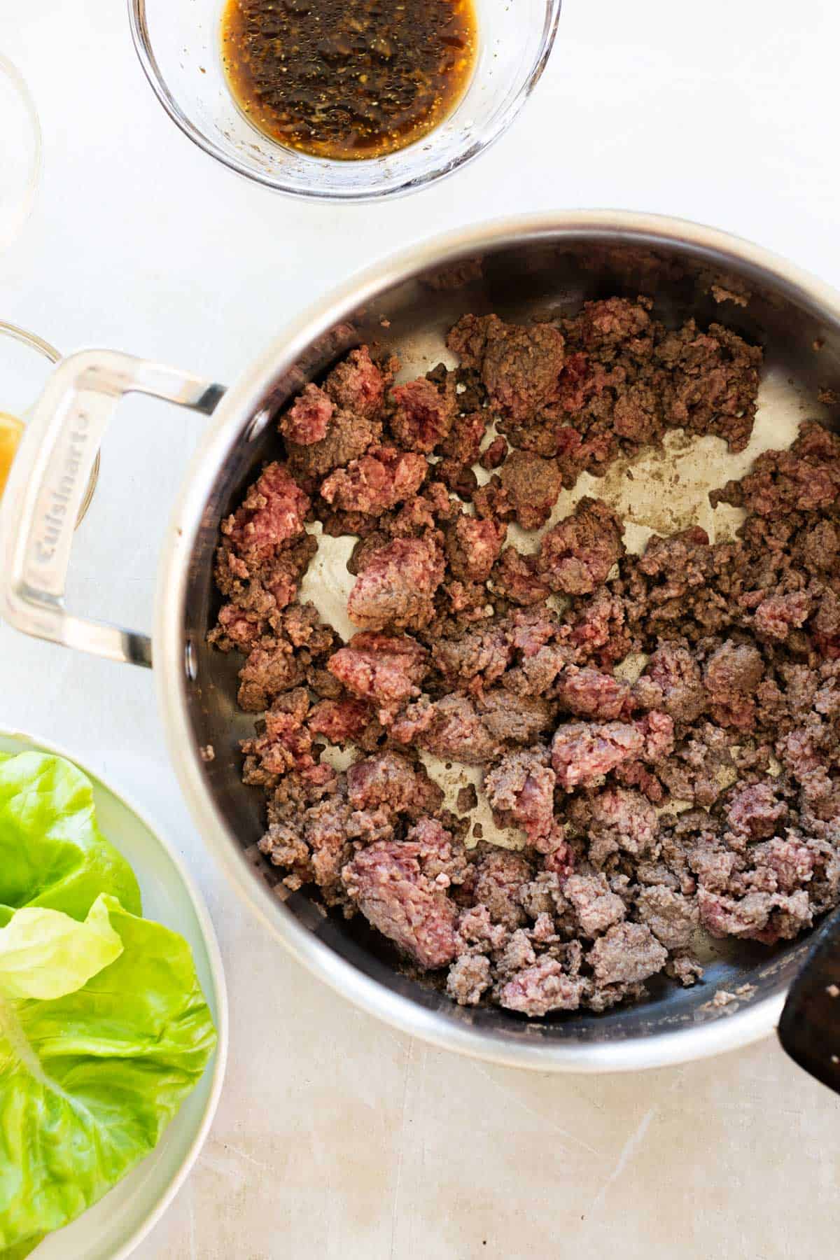 Ground beef cooking in a stainless steel saucepan, with a bowl of sauce above and a plate of green lettuce leaves to the side on a white surface.
