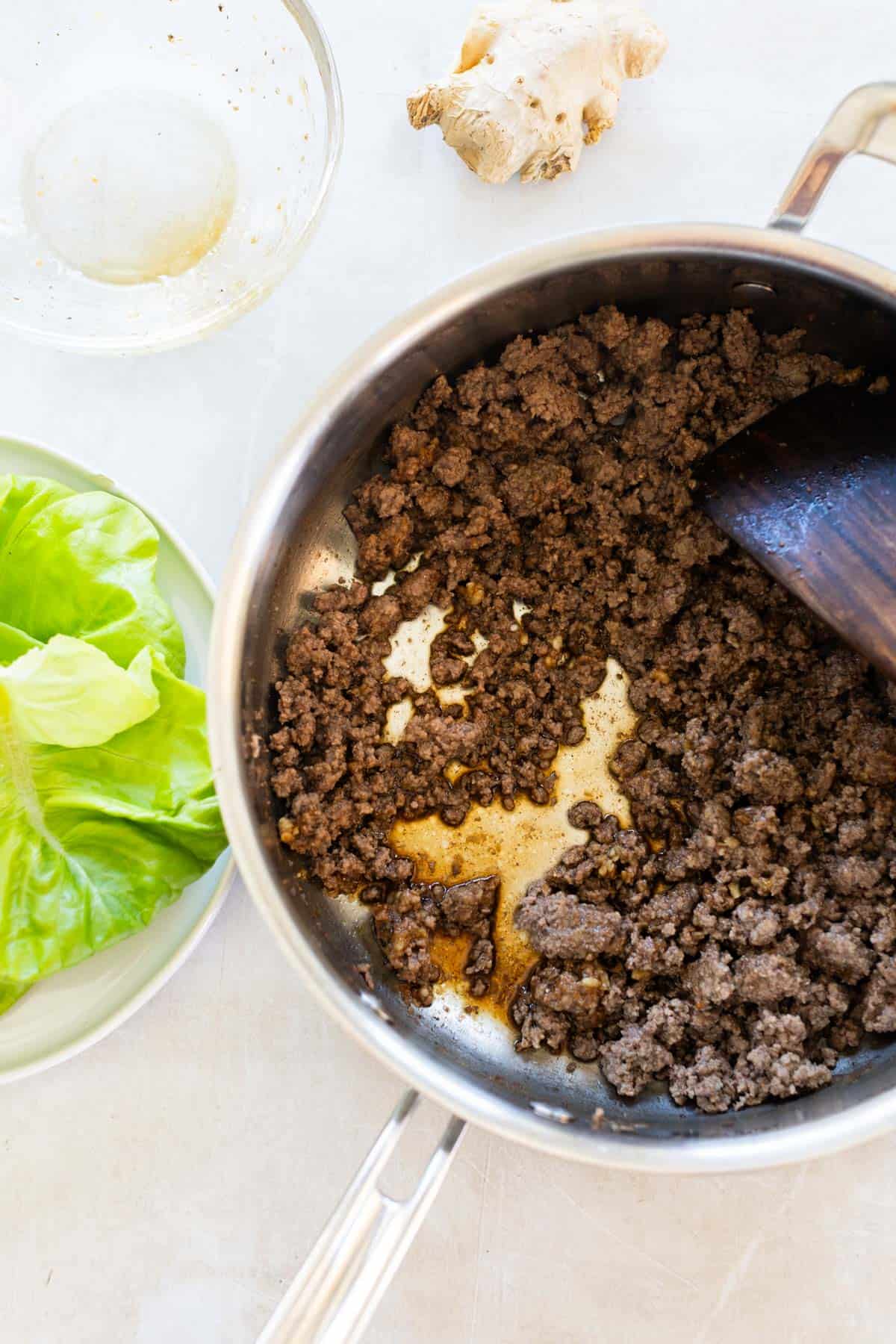 Cooked ground beef in a saucepan with a wooden spatula, fresh lettuce leaves on a green plate, and a piece of ginger and empty bowl nearby.